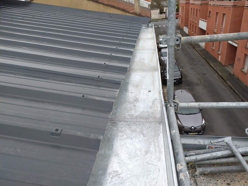Toiture en bac acier gris avec chéneau en zinc installé sur un chantier sécurisé par échafaudage.
