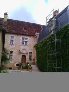 Manoir ancien en pierre avec échafaudage, mur de lierre et porte sculptée décorée de fleurs.