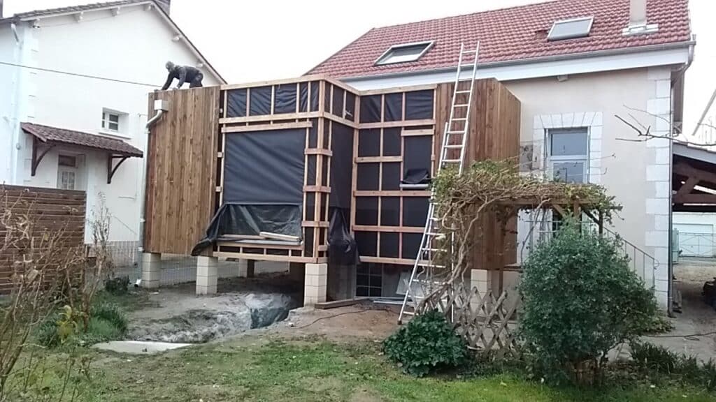 Extension en bois : chantier de construction et rénovation Extension de maison en ossature bois en chantier avec ouvrier sur le toit, membrane d'étanchéité et jardin.