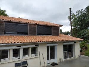 Maison moderne à étage avec toiture en tuiles orange, volets roulants gris et terrasse sous un ciel couvert.