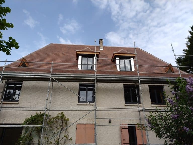 Maison en rénovation avec échafaudage devant une façade beige et toiture en tuiles brunes sous un ciel bleu.
