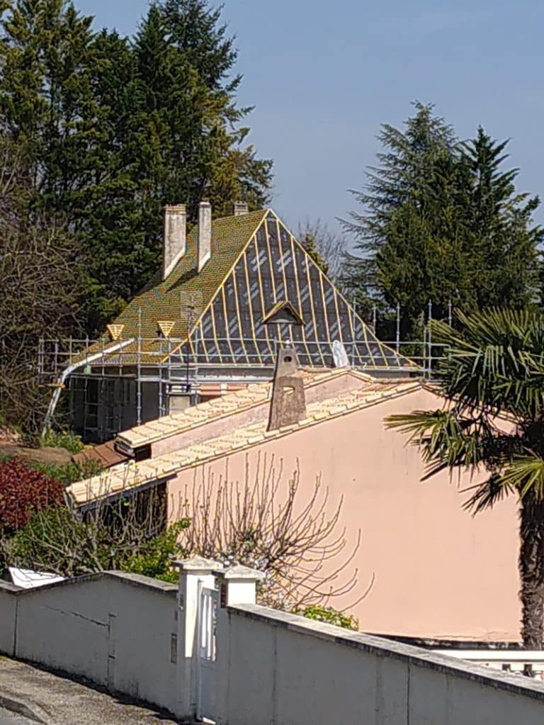 Rénovation de toiture avec échafaudage et isolant noir sur une maison ancienne, devant une bâtisse rose.