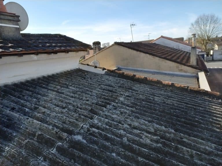 Toiture en plaques ondulées grises usées avec cheminée et antenne parabolique sous un ciel bleu.